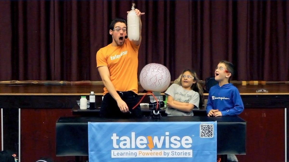 Science Hero holds up some cool science stuff while kids look on in amazement.