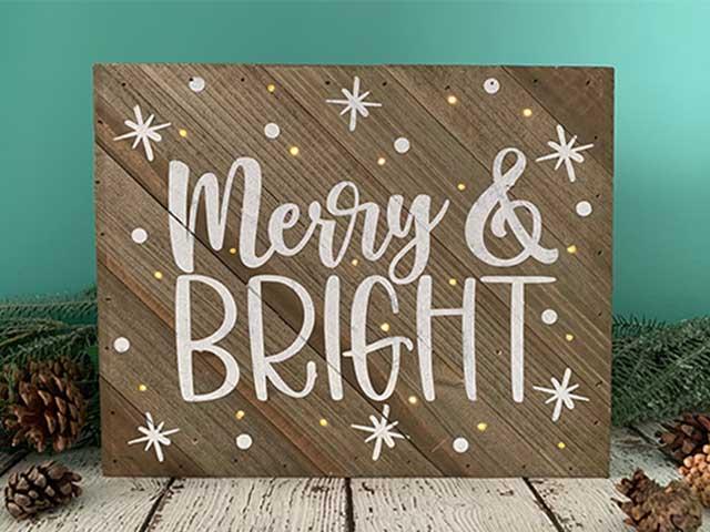 A wooden sign says "Merry & Bright" in white letters surrounded by small lights and snowflakes.